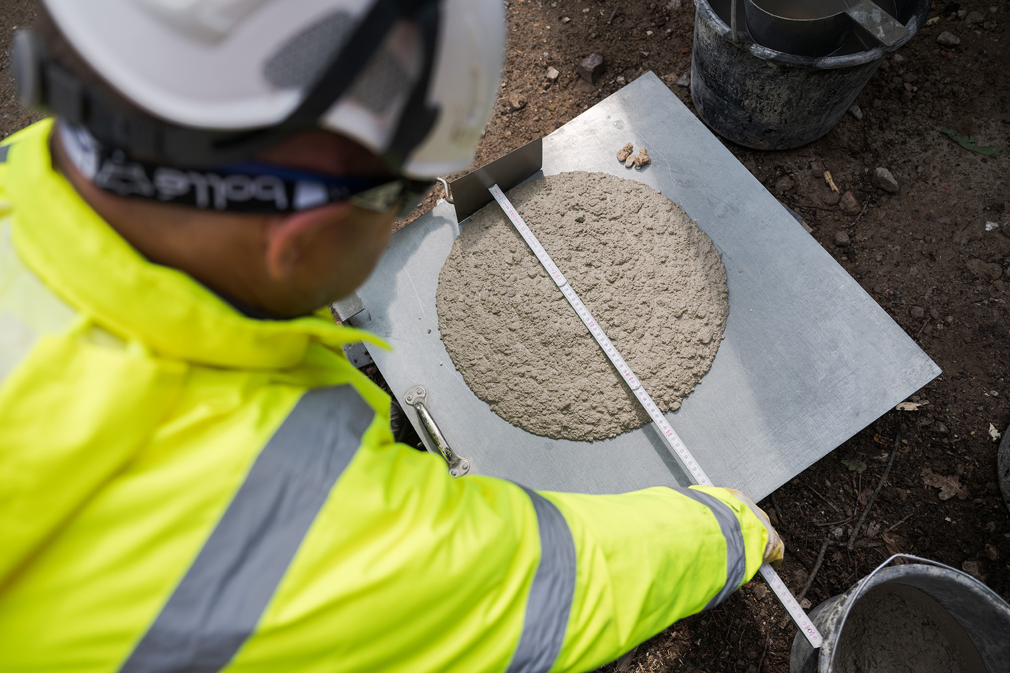 Ein Bauarbeiter in gelber PSA und Schutzhelm misst die Ausbreitung von Frischbeton auf einer Metallplatte mit einem Lineal, durchgeführt im Freien.