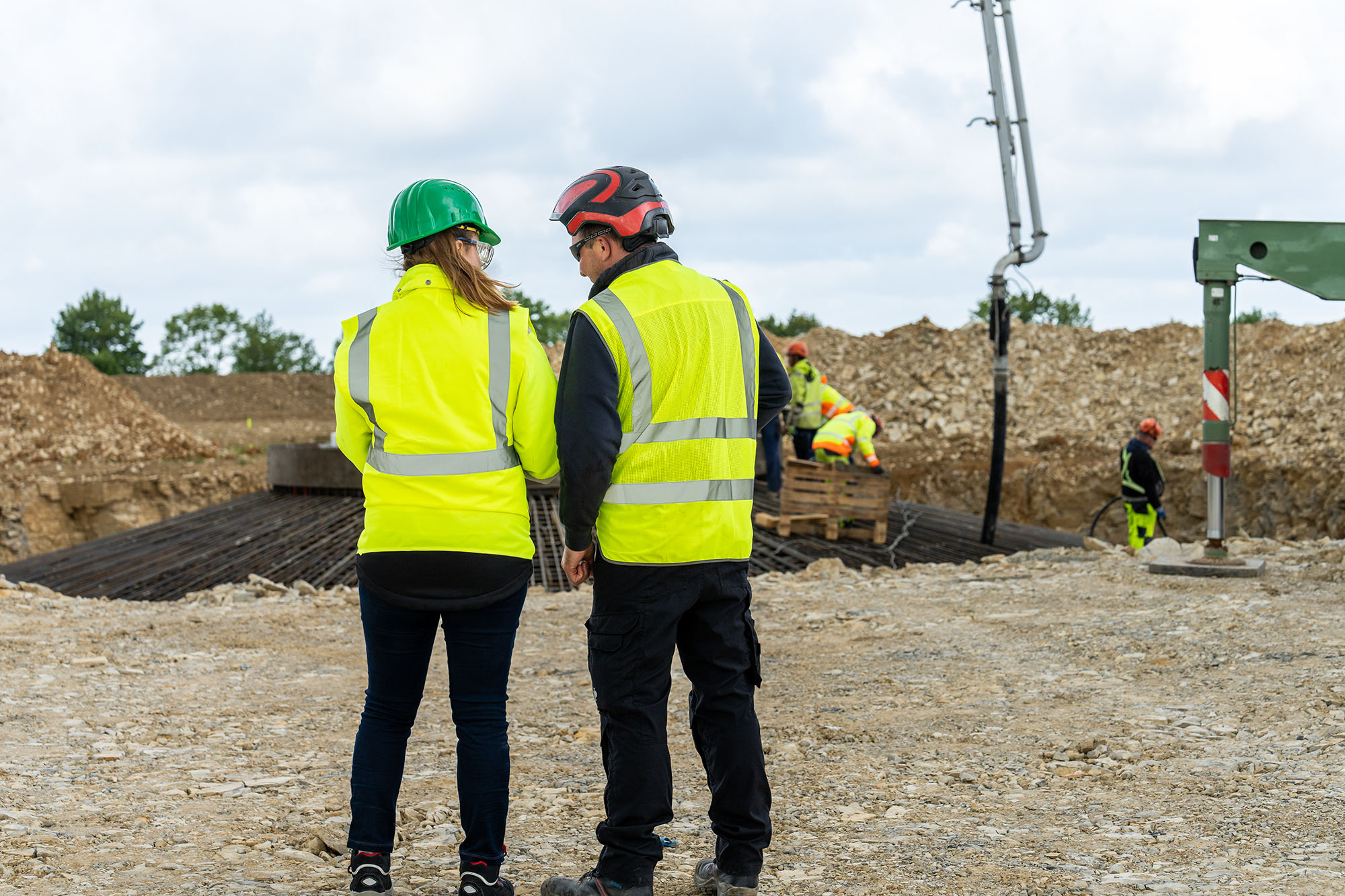 Zwei Mitarbeiter in Warnwesten und Helmen stehen auf einer Baustelle und betrachten die Arbeiten. Im Hintergrund sind eine Betonpumpe, Erdaufschüttungen und weitere Arbeiter in PSA zu sehen.