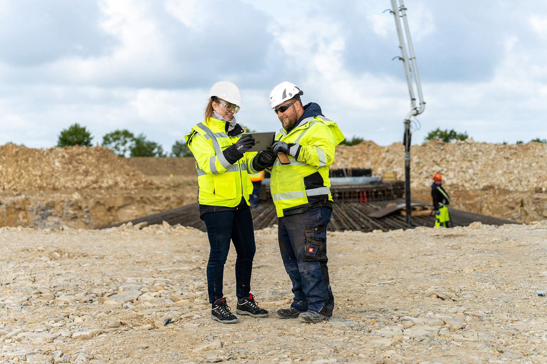 Zwei Mitarbeiter in Warnwesten und Helmen stehen auf einer Baustelle und betrachten die Arbeiten. Im Hintergrund sind eine Betonpumpe, Erdaufschüttungen und weitere Arbeiter in PSA zu sehen.