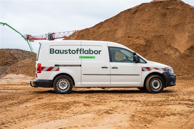 Weißer Lieferwagen mit der Aufschrift "Baustofflabor" auf sandigem Untergrund vor einem großen Erdhaufen; im Hintergrund sind Teile eines Krans zu sehen.
