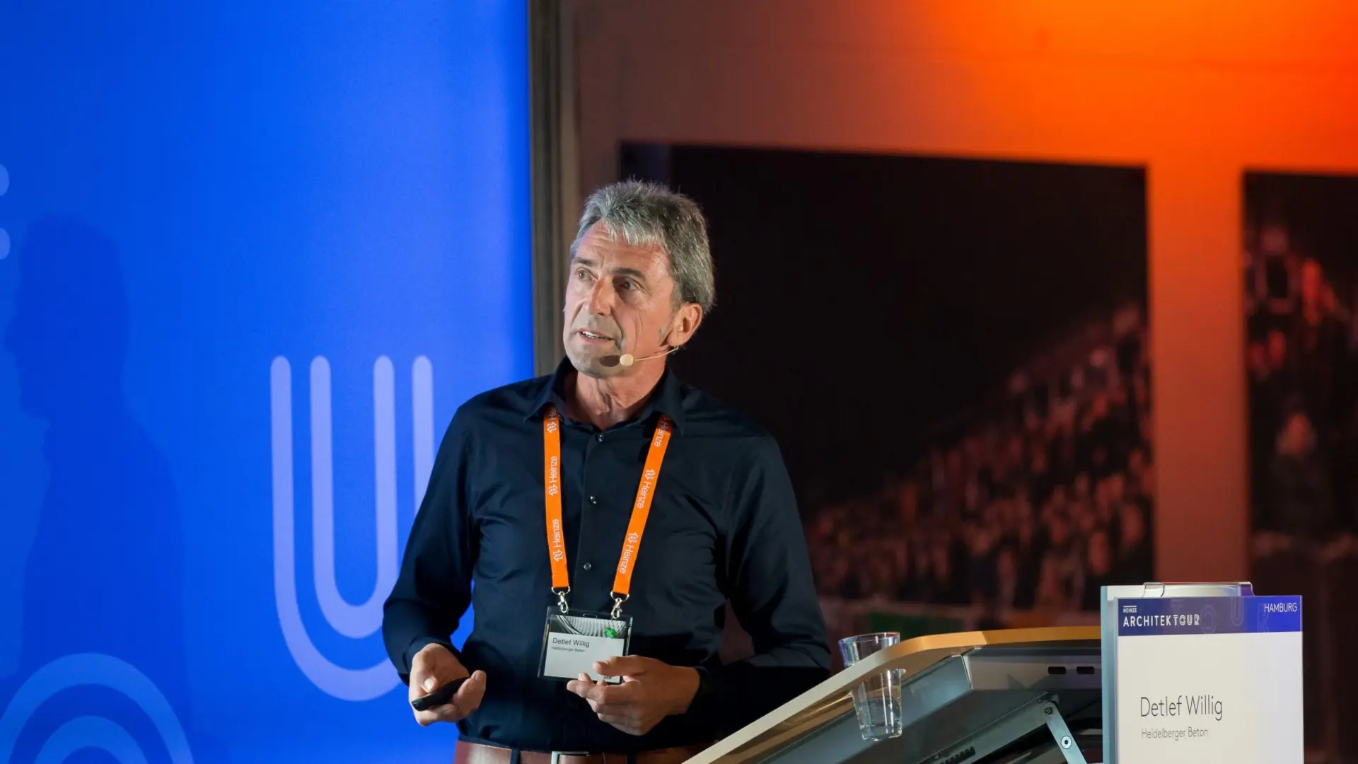 Ein Experte der Betotech steht vor einem blauen "Heinze ArchitekTOUR"-Banner, hält einen Vortrag und trägt ein Namensschild mit orangener Lanyard. Rechts im Vordergrund ist ein Rednerpult sichtbar.