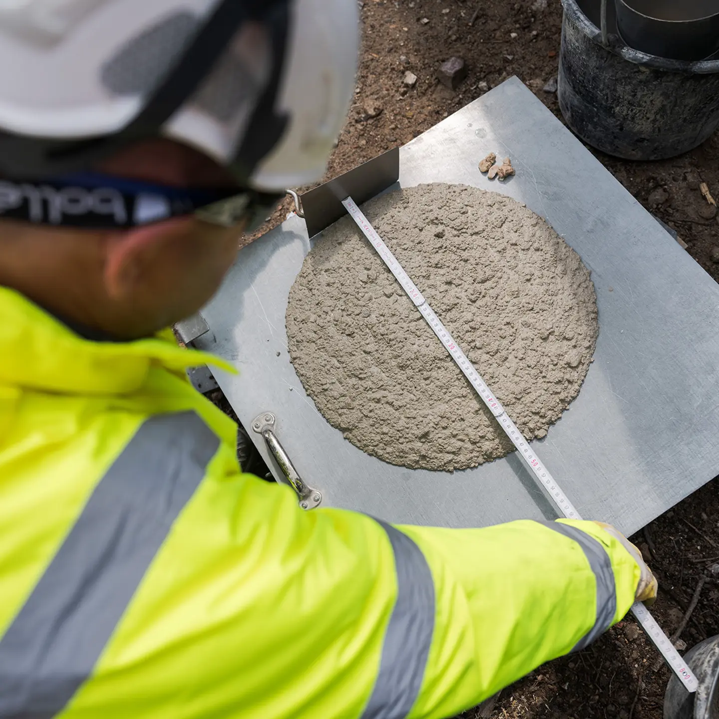 Ein Bauarbeiter in gelber PSA und Schutzhelm misst die Ausbreitung von Frischbeton auf einer Metallplatte mit einem Lineal, durchgeführt im Freien.