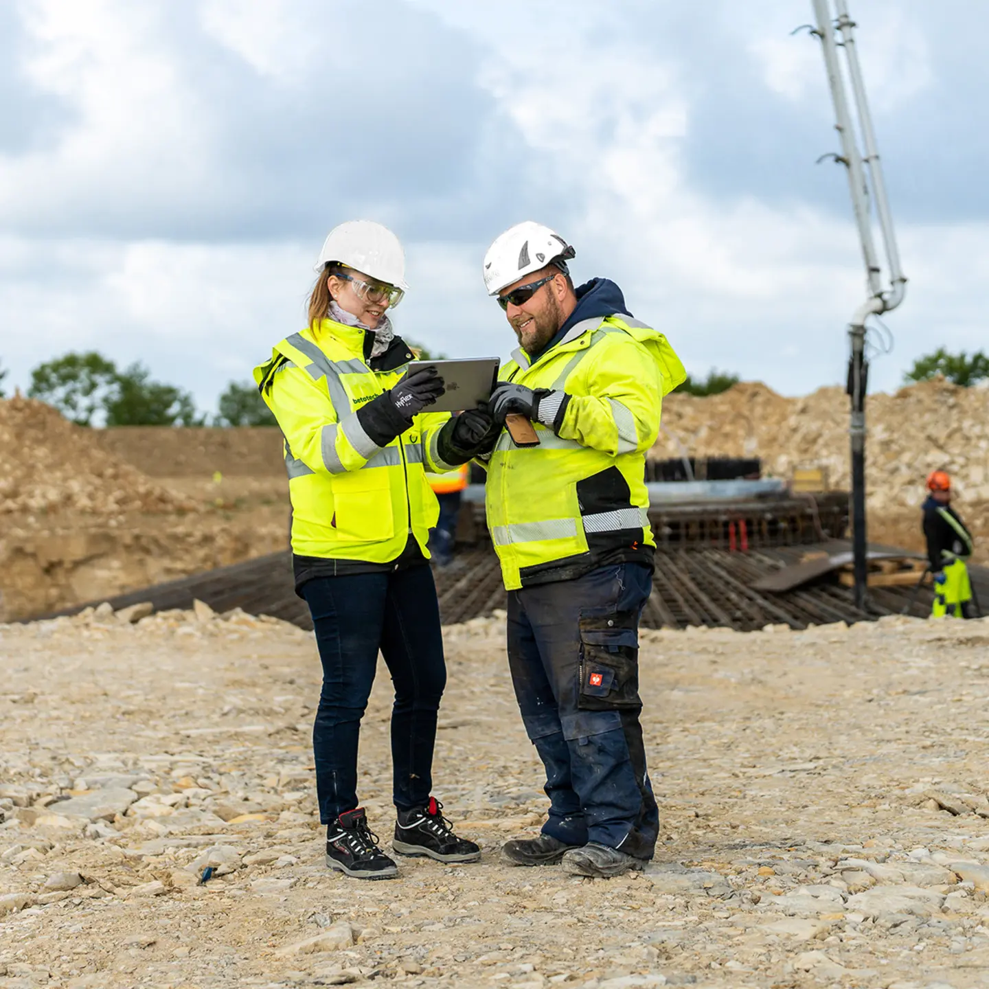 Zwei Mitarbeiter in Warnwesten und Helmen stehen auf einer Baustelle und betrachten die Arbeiten. Im Hintergrund sind eine Betonpumpe, Erdaufschüttungen und weitere Arbeiter in PSA zu sehen.