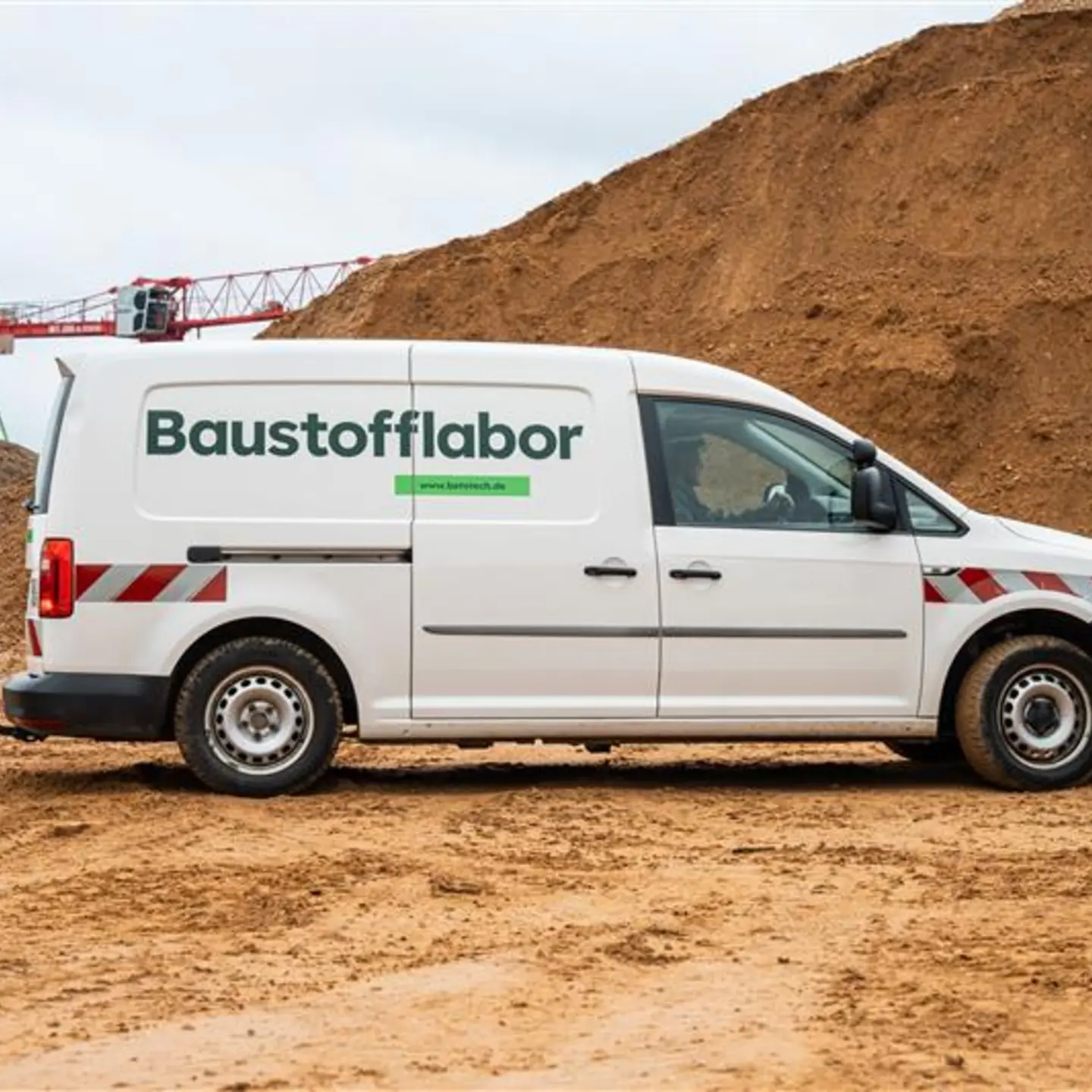 Weißer Lieferwagen mit der Aufschrift "Baustofflabor" auf sandigem Untergrund vor einem großen Erdhaufen; im Hintergrund sind Teile eines Krans zu sehen.