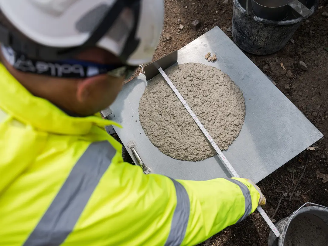 Ein Bauarbeiter in gelber PSA und Schutzhelm misst die Ausbreitung von Frischbeton auf einer Metallplatte mit einem Lineal, durchgeführt im Freien.