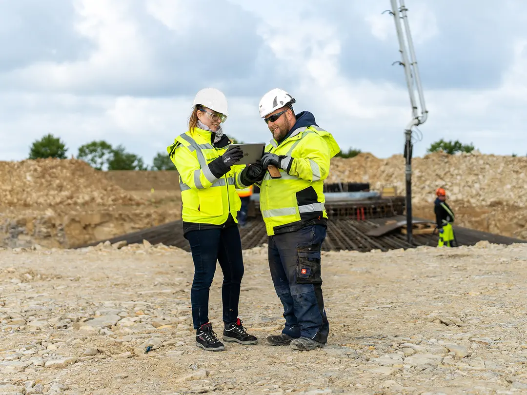 Zwei Mitarbeiter in Warnwesten und Helmen stehen auf einer Baustelle und betrachten die Arbeiten. Im Hintergrund sind eine Betonpumpe, Erdaufschüttungen und weitere Arbeiter in PSA zu sehen.