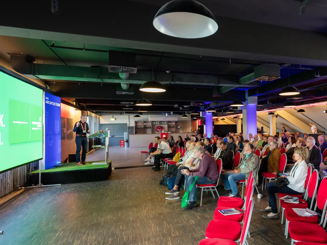 Ein Experte der Betotech steht auf einer Bühne links, neben einer großen Präsentationsleinwand, während ein Publikum in roten Stühlen rechts sitzt und zuhört.