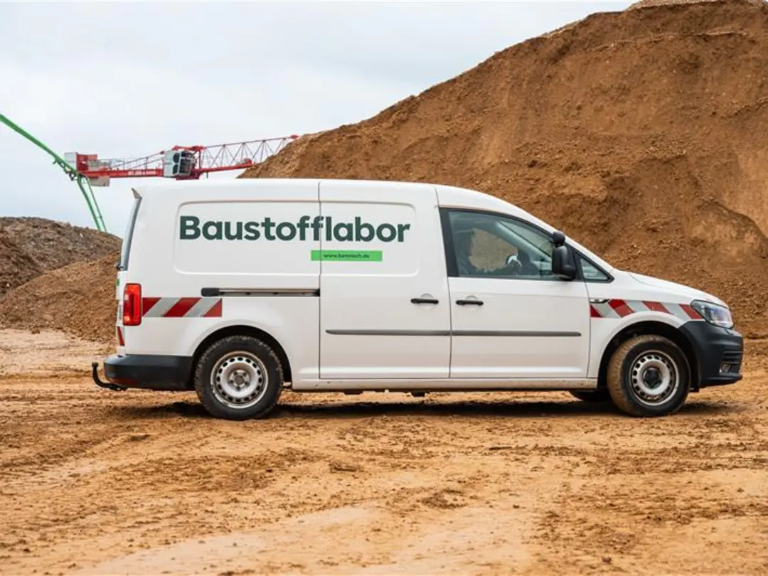 Weißer Lieferwagen mit der Aufschrift "Baustofflabor" auf sandigem Untergrund vor einem großen Erdhaufen; im Hintergrund sind Teile eines Krans zu sehen.